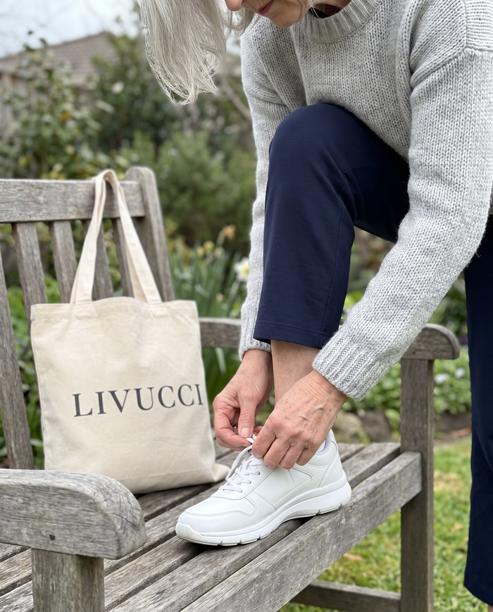 Woman on a garden bench lacing a white walking sneaker beside a Livucci tote